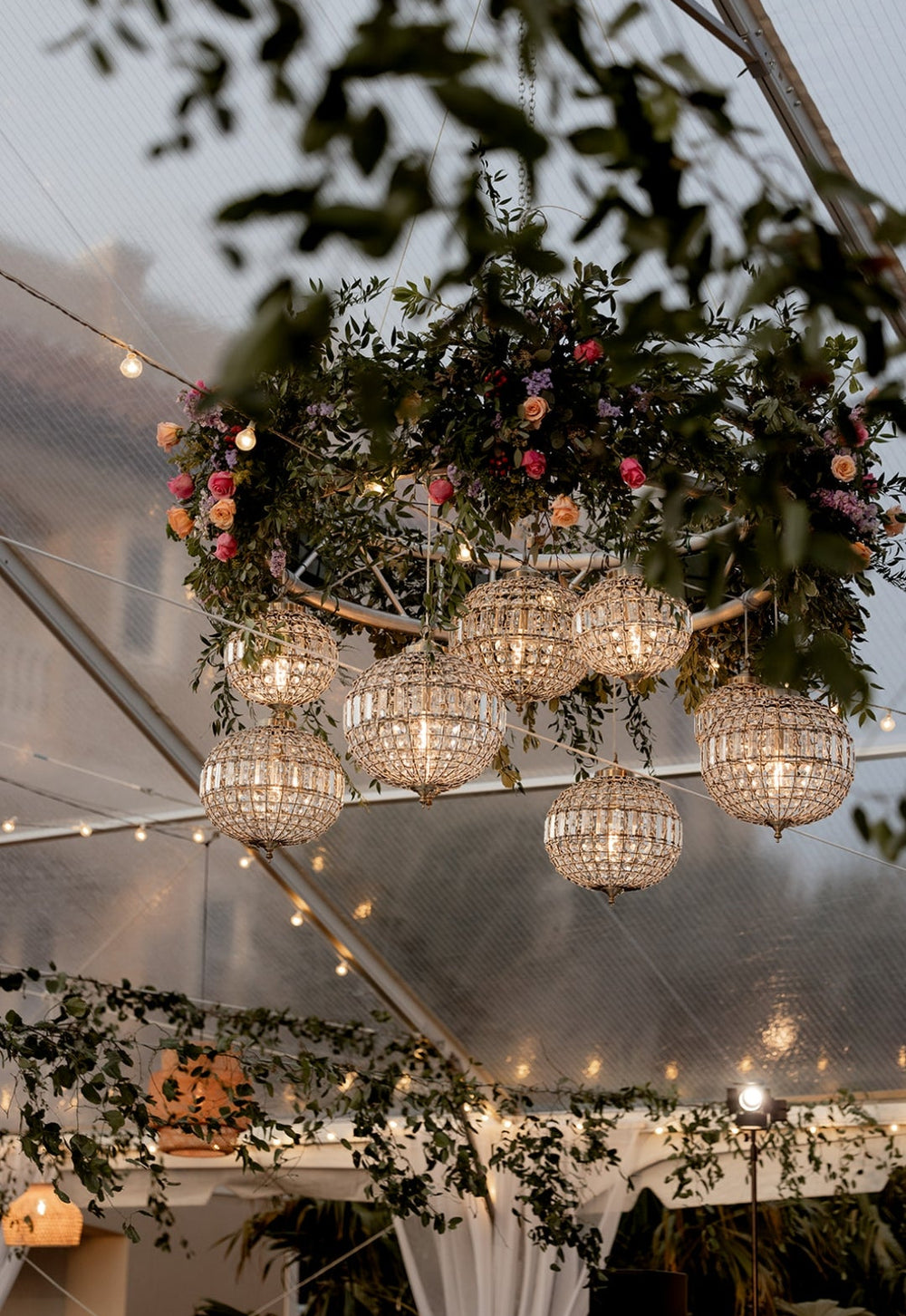 Decorative chandelier with hanging lights and floral arrangements inside a tented structure.