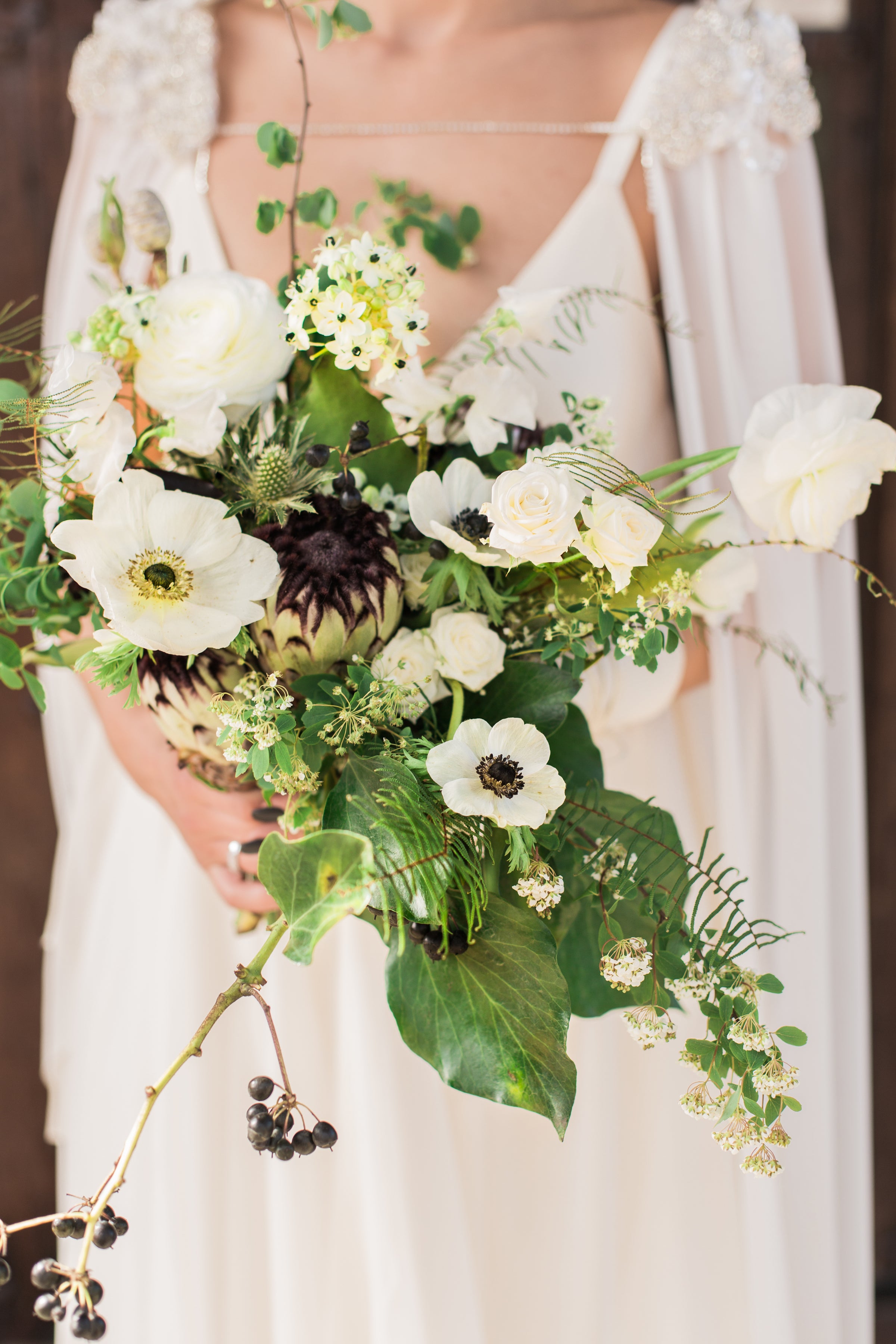 A garden style bridal bouquet. A variety of greenery, Flowers are white with dark centers. Focal flowers are white king protea and panda white anemones.