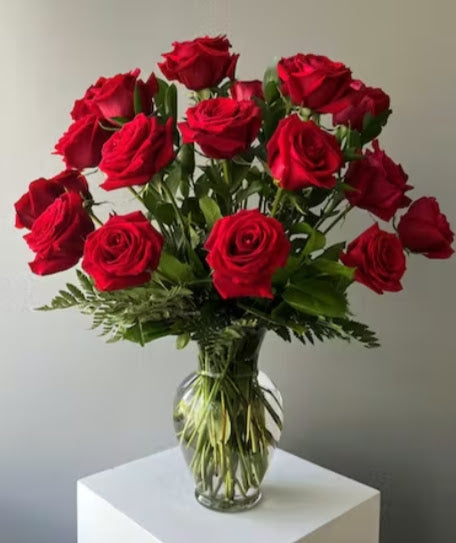 24 long stem roses with lush greenery arranged in a clear glass vase.